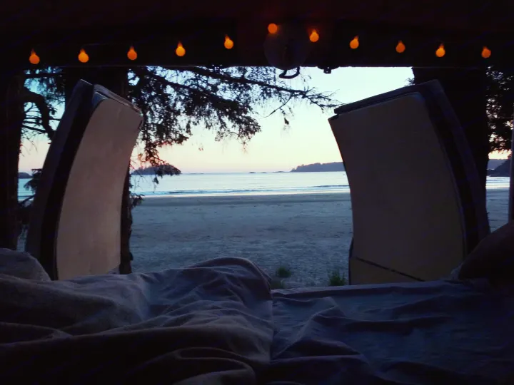 Cozy rv rental bed view of a serene beach sunset. String lights glow inside the van, framing the tranquil ocean and distant shore.. Ford Econoline 2006