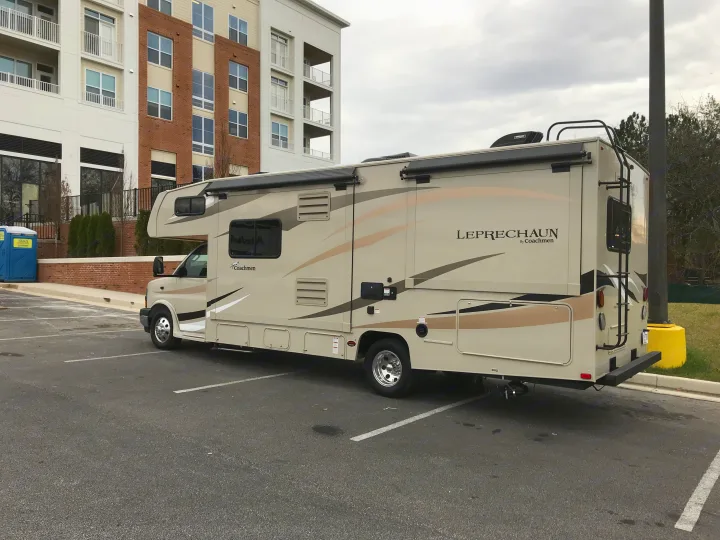 Tan Coachmen Leprechaun Class C rv rental parked in a lot with a modern building in the background, ready for travel.. Coachmen Leprechaun 2018