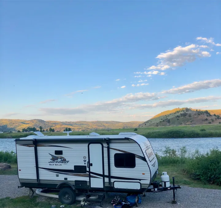 A white Jayco travel trailer (RV rental) parked on gravel next to a river with green hills & a blue sky. Ideal for nature lovers.. Jayco Other 2020