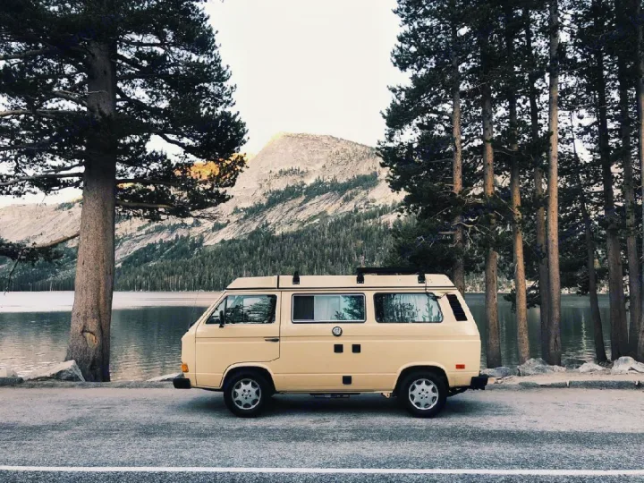 An RV rental, a classic yellow camper van, parked beside a tranquil mountain lake and forest pines.. Volkswagen Westfalia 1982