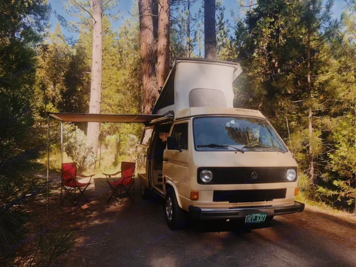 A beige VW Westfalia camper van, pop-top extended, with awning and chairs, parked on a forest road. Ideal RV rental.. Volkswagen Westfalia 1982