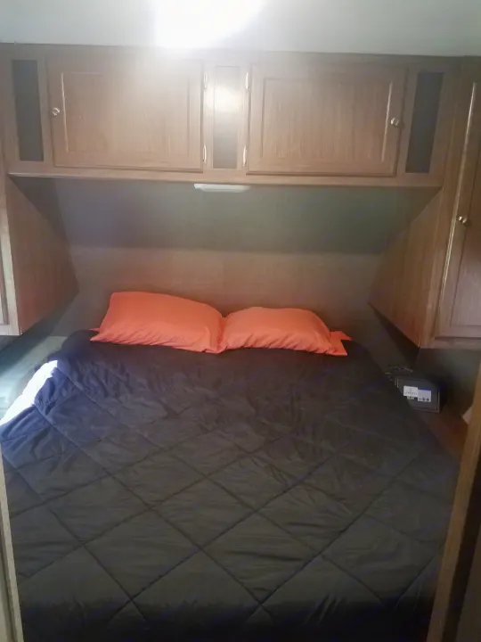Compact RV rental bedroom featuring a comfy bed with orange pillows and built-in overhead cabinets, ideal for road trips.. Heartland Pioneer 2016