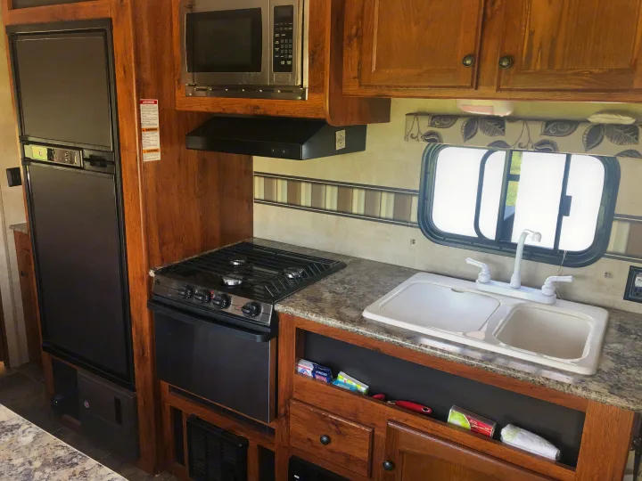 This RV rental kitchen features a refrigerator, microwave, stove, oven, and double sink with wooden cabinets.. Keystone Hideout 2016