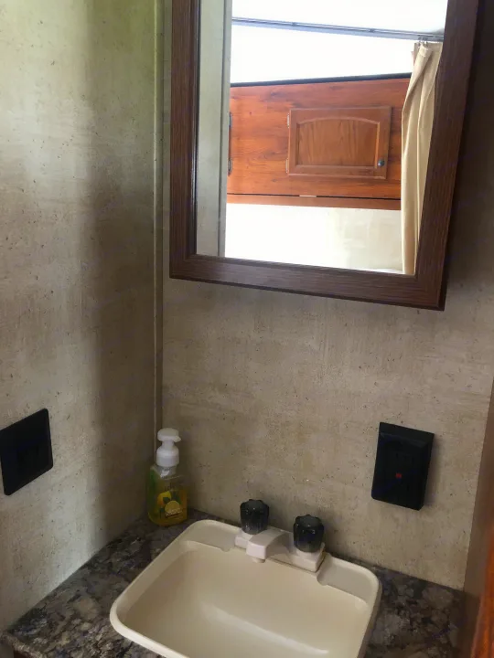 Compact RV rental bathroom features a sink, soap, and mirror reflecting a wood cabinet & window. Textured walls & dark counter.. Keystone Hideout 2016