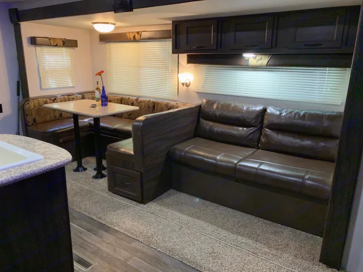 RV rental interior: A dark U-shaped dinette & brown sofa, with overhead cabinets, blinds, and a mix of wood & carpet flooring.. Pacific Coachworks Tango 2020
