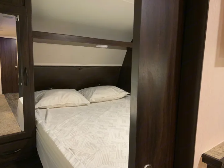 Cozy RV rental bedroom featuring a comfortable bed with white bedding, dark wood cabinets, and built-in shelving.. Pacific Coachworks Tango 2020