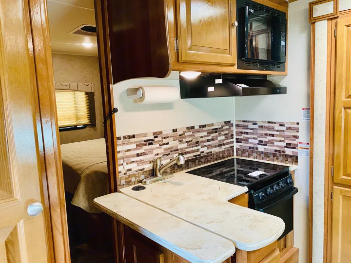RV rental: Compact kitchen with sink, stovetop, microwave, tiled backsplash, and bedroom view through an open door.. Coachmen Leprechaun 2018