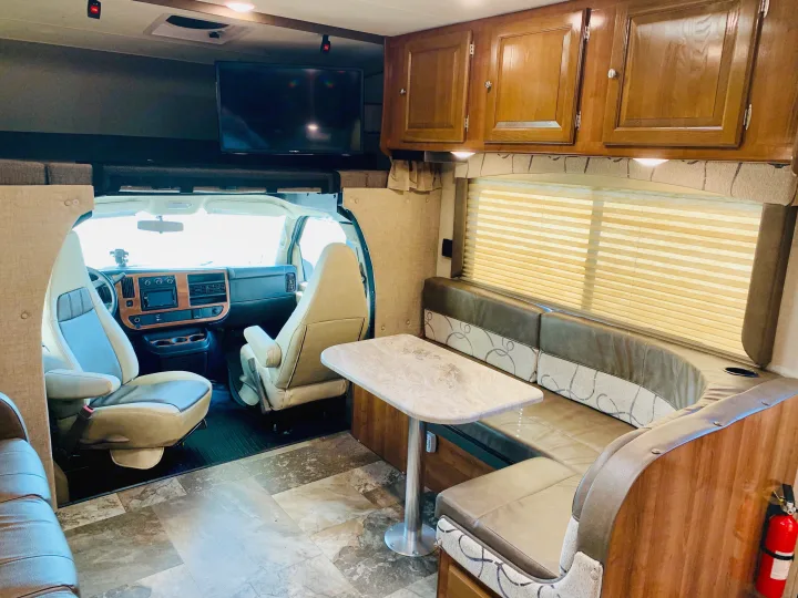 Bright motorhome RV rental interior: view of dining booth, driver's cabin, overhead bed, and mounted TV. Tiled floor.. Coachmen Leprechaun 2018