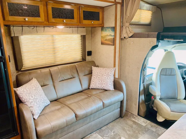 Interior of an RV rental showing a brown sofa with decorative pillows, wooden cabinets, and the front driving area.. Coachmen Leprechaun 2018
