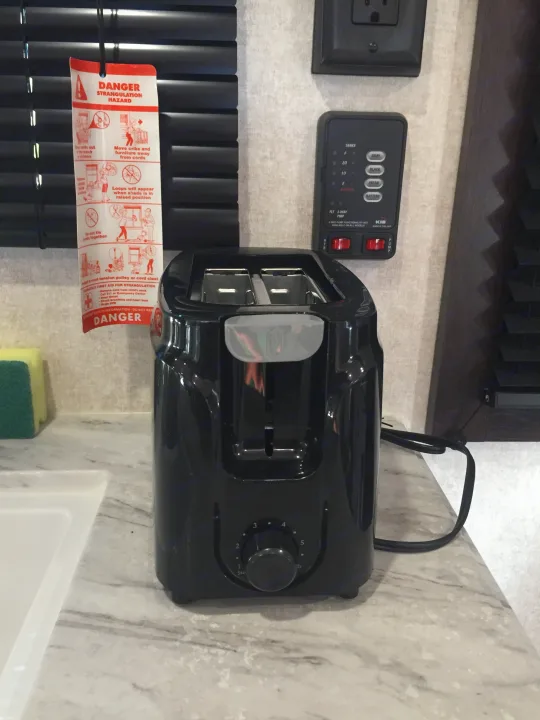 RV rental interior view: Black toaster, RV tank monitor, power outlet, and window blinds with a safety warning tag.. Jayco Jay Flight SLX 2019