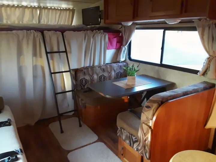 RV rental interior: a cozy dinette with patterned seats, an overhead bunk bed with ladder & privacy curtains, plus a TV.. Coachmen Freelander 2011