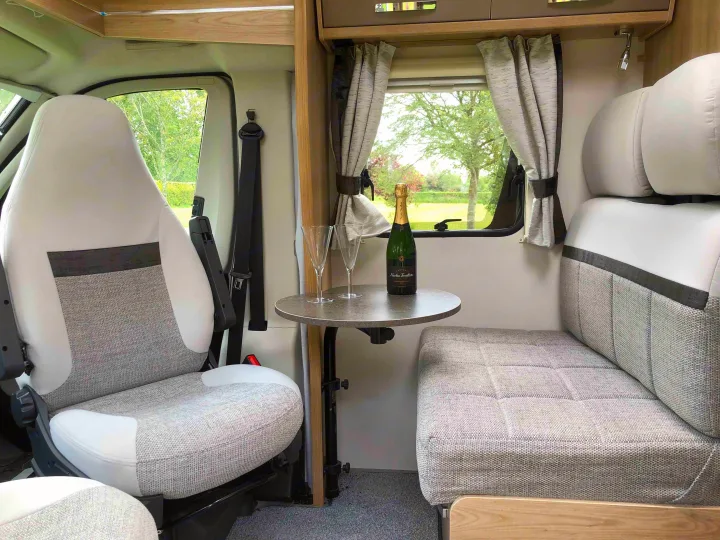 Cozy RV rental interior with champagne, showing a swivel seat, bench, and table by a window with a green view.. Elddis Accordo 135 2019