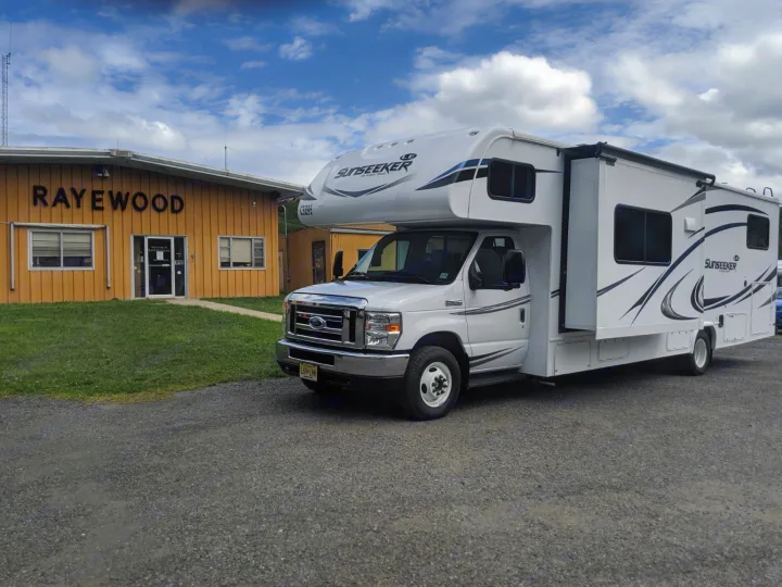 A white Sunseeker Class C RV rental, a recreational vehicle, parked on gravel in front of the yellow Rayewood building.. Forest River Sunseeker 2025