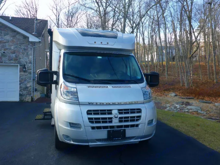 Front view of a silver Winnebago RV rental on a paved driveway, a stone house and winter trees behind it.. Winnebago Trend 2016