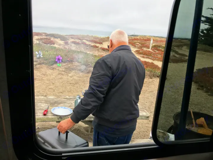 RV rental: Man grilling at a coastal campsite, viewed from inside. Ocean & decorative flowers in view.. Jayco Jay Flight SLX 2017