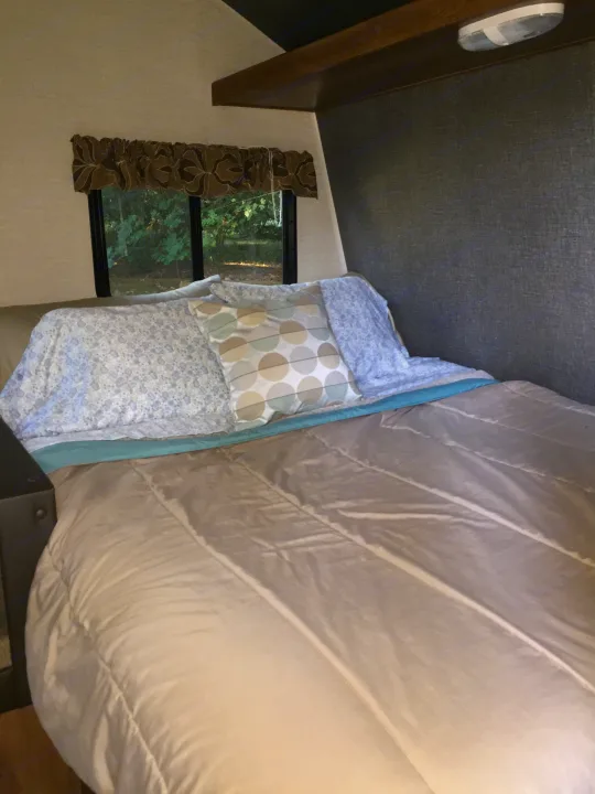 Inside a comfortable RV rental bedroom, featuring a bed with patterned pillows and a window overlooking lush green nature.. Keystone RV Summerland Mini 2015