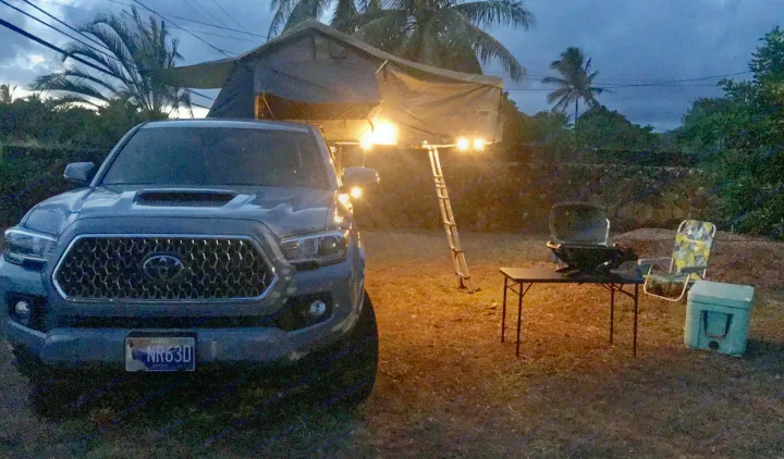 A grey Tacoma truck with a lit rooftop tent & ladder. A grill, table, and cooler are set up for camping. Ready for an RV rental adventure!. Other Other 2019