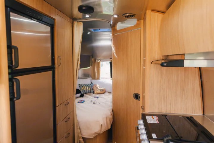 Interior of a modern RV rental: a Dometic fridge & kitchen flow into a cozy bedroom with a comfortable bed. Amenities include wood cabinetry.. Airstream Flying Cloud 2020