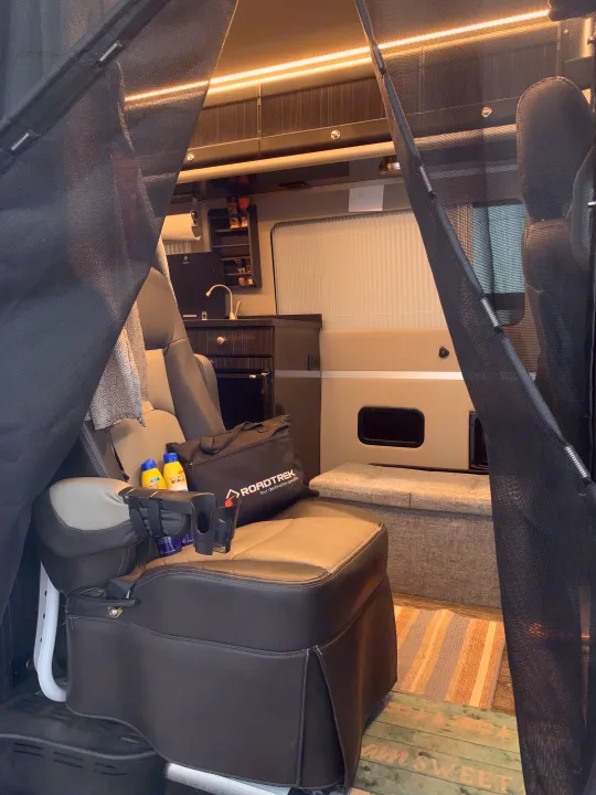 Inside an RV rental: a plush seat with a 'Roadtrek' bag and cupholders, a compact kitchen, and black mesh privacy screens.. Roadtrek RS E-Trek 2018