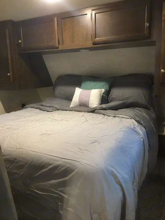 Compact RV rental bedroom: comfy bed with grey bedding, accent pillows, and dark wood overhead cabinets.. Starcraft Autumn Ridge 2018