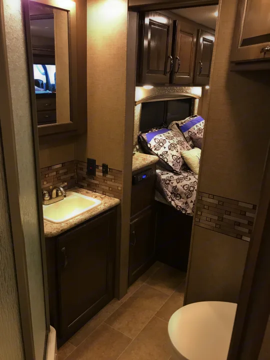 Inside this RV rental: a modern bathroom with sink & stylish tile, leading to a cozy bedroom with patterned bedding & dark wood cabinets.. Thor Chateau 2020