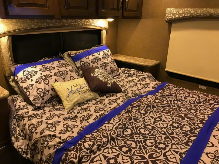 RV rental bed with intricate brown and white patterned bedding, blue accents, overhead cabinets, and a window shade.. Thor Chateau 2020