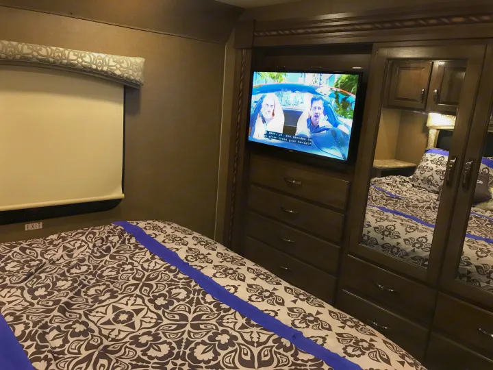 Cozy RV rental bedroom with comfortable, patterned bed, TV showing a film, and dark wood built-in storage cabinetry.. Thor Chateau 2020