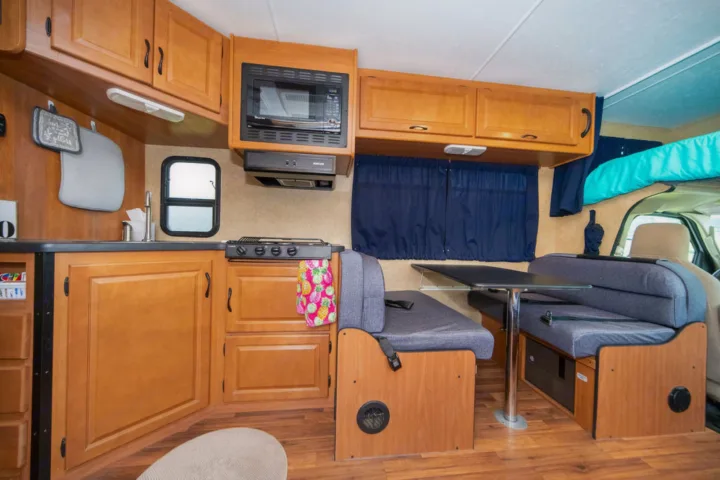 RV rental interior with a compact kitchen, dining booth, and bed above the cab, featuring wood cabinetry.. Other Other 2015