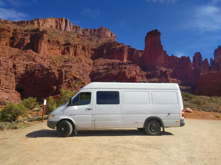 White RV rental van on a dirt road, backed by towering red rock cliffs under a clear blue sky. Ideal for desert road trips.. Mercedes-Benz Sprinter 2005