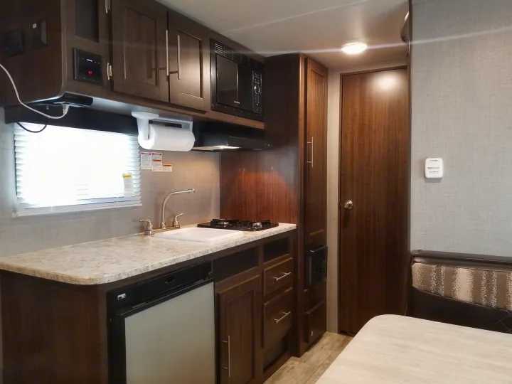 RV rental kitchen with sink, stove, microwave, and fridge. Dark wood cabinets, light countertops, and a dining area are visible.. Crossroads RV Zinger Lite 2019
