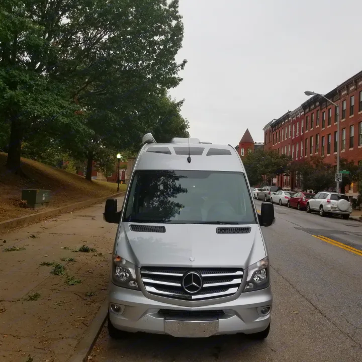 Silver Mercedes-Benz Sprinter RV rental van parked on a city street. Lush trees line the sidewalk, with brick buildings in the background.. Roadtrek Ssagile 2014