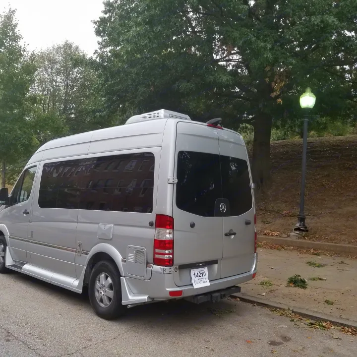 A silver Mercedes Sprinter RV rental van with a temporary tag, parked on a street. Trees & a lit lamp in background.. Roadtrek Ssagile 2014