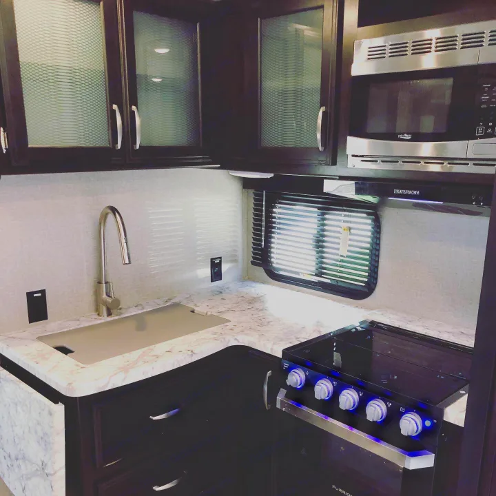 Modern RV rental kitchen with dark cabinets, marble counters, microwave, and stove with blue light. Ready for travel.. Grand Design Imagine 2019