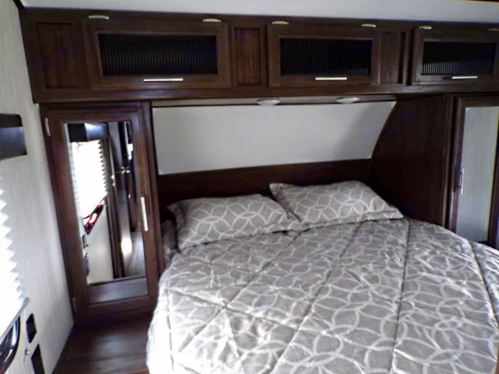 Cozy RV rental bedroom with a patterned bed, dark wood overhead storage, and mirrored wardrobes for comfortable travel.. Forest River Alpha Wolf 2020