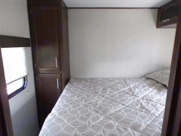 RV rental bedroom shows a bed with grey/white comforter, a dark wood cabinet, and window blinds. Compact and functional space.. Forest River Alpha Wolf 2020