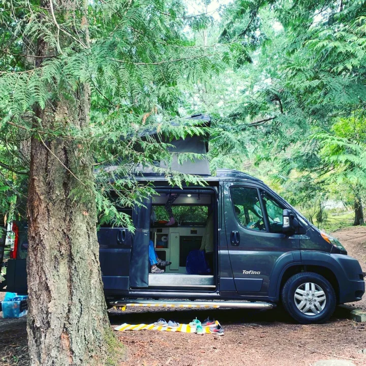 A grey 'rv rental' camper van, door open with shoes neatly outside, is parked among lush forest trees for camping.. PleasureWay Tofino 2019