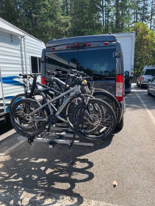 Dark gray RV rental van with a bike rack holding multiple mountain bikes, parked next to a white travel trailer.. PleasureWay Tofino 2019