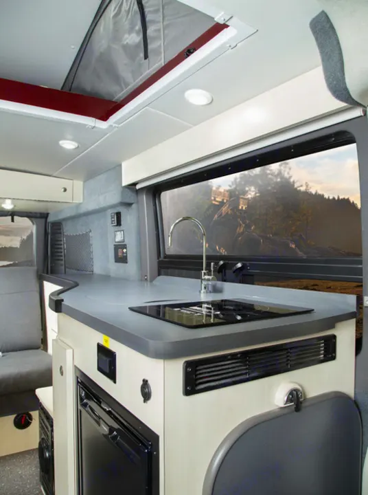 Modern RV rental kitchen with sink, stove, and fridge inside a camper van, offering a scenic window view.. PleasureWay Tofino 2019