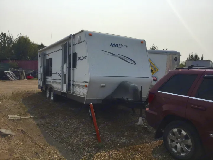 This white Maxlite travel trailer, a potential RV rental, sits on a gravel lot beside a red SUV.. Max Lite ML26RK 2006