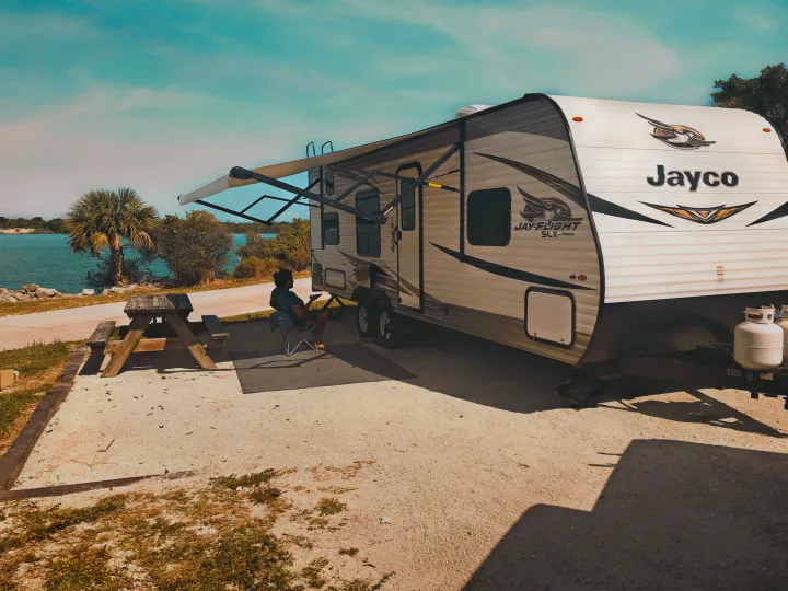 A person relaxes under the awning of a Jayco RV rental, parked seaside with a picnic table.. Jayco Jay Flight SLX 2019