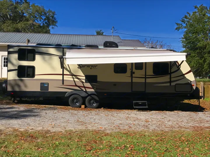 A large beige and brown 'Surveyor' travel trailer with an extended awning, ideal for rv rental, is parked on a gravel driveway.. Forest River Surveyor 2015