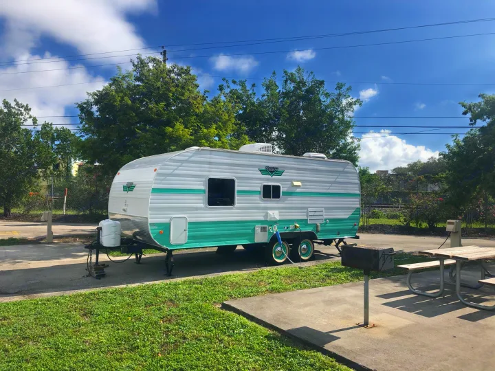 A charming white and teal Retro RV rental trailer parked at a sunny campsite with a picnic table and grill.. Riverside Rv 189R Retro 2018