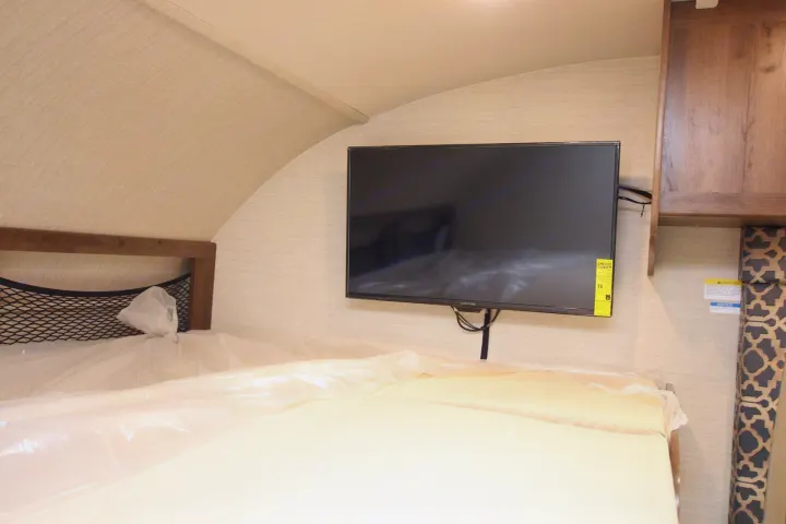 Modern RV rental bedroom with a bed featuring mesh storage, a wall-mounted TV, and wooden overhead cabinets.. Jayco Redhawk 2019
