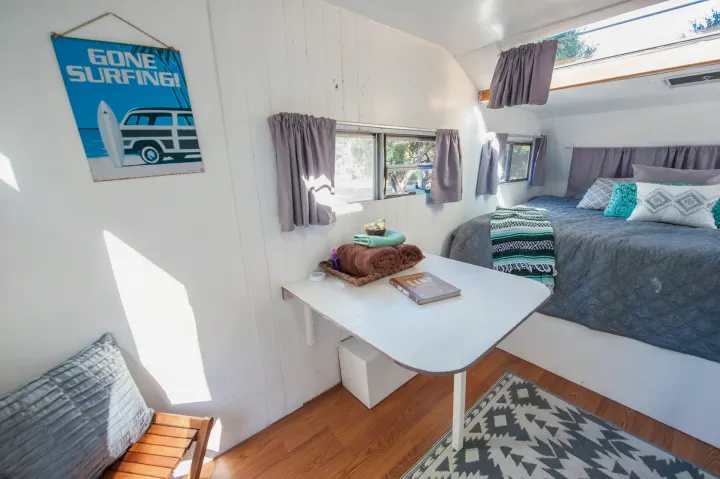 Bright rv rental interior with a comfy bed, decorative pillows, a table with towels & a book, and a "Gone Surfing" sign.. Custom Camper Trailer 1973