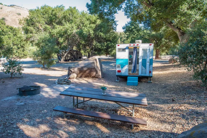 This image shows a colorful blue and white rv rental at a serene campsite with a picnic table and fire pit, surrounded by trees.. Custom Camper Trailer 1973