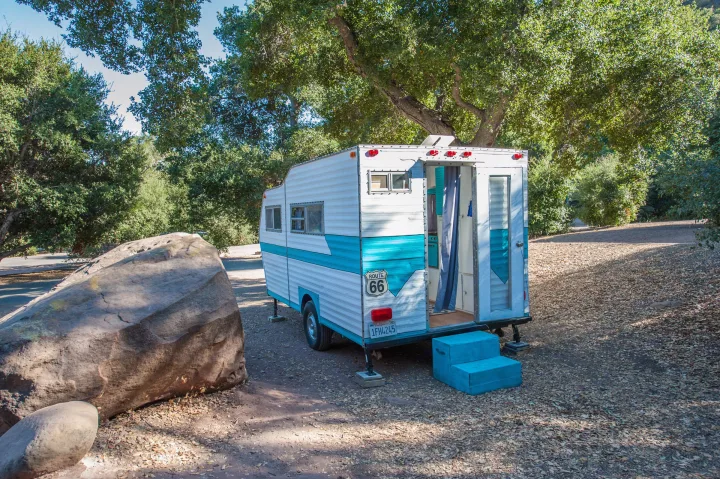 Charming vintage turquoise RV rental with a Route 66 sign, parked under trees. Door open, ready for adventure.. Custom Camper Trailer 1973