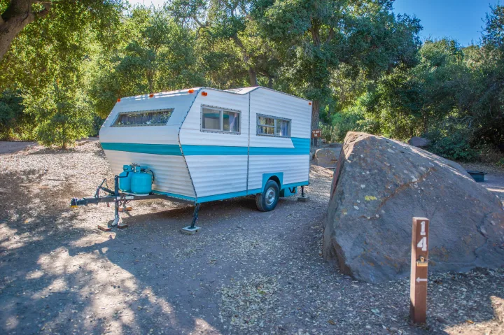 A classic white and teal RV rental is settled into campsite 14, shaded by trees next to a large rock.. Custom Camper Trailer 1973