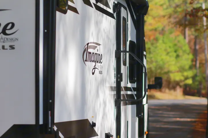 Side of a white Grand Design Imagine XLS RV with black graphics & door handle, parked outdoors. Great for an rv rental experience!. Grand Design Imagine XLS 19BWE 2020
