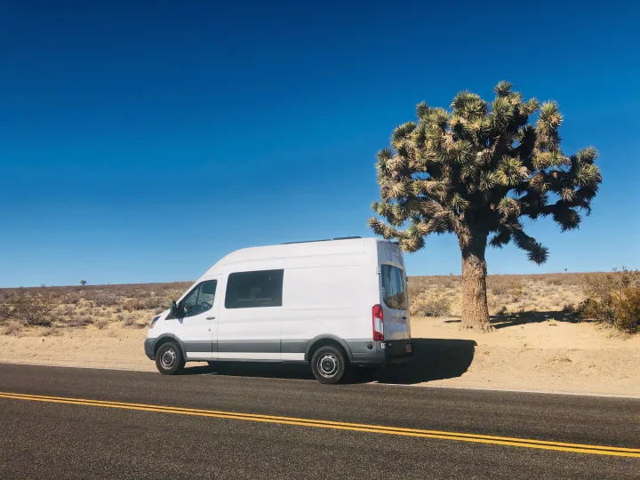 White RV rental van parked beside a Joshua tree on a desert road under a clear blue sky, ideal for a road trip.. Ford Transit 2015