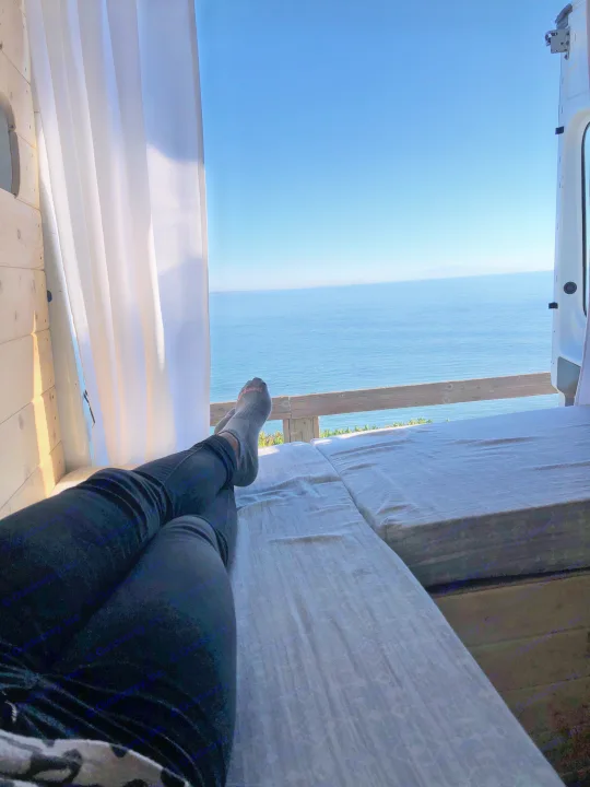 Enjoying the stunning ocean view from an RV rental! Legs stretched out on the bed, looking out at the blue sea.. Ford Transit 2015